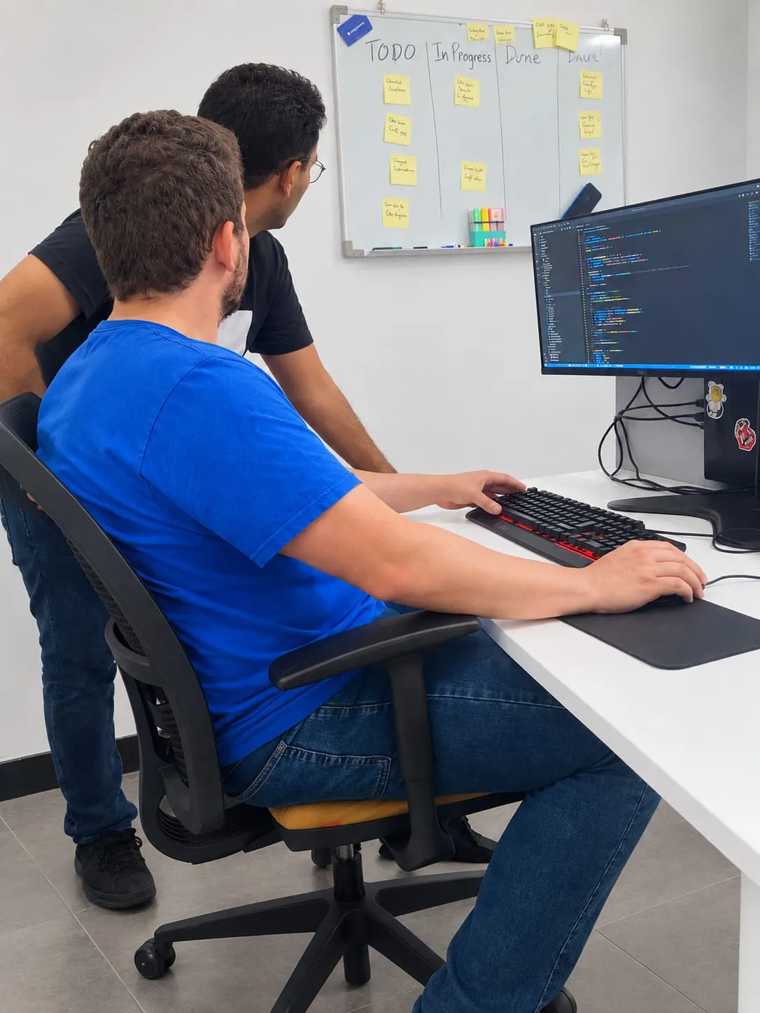 Two people at a desk reading the same screen of code, one at the keyboard, a board of sticky notes on the wall behind them.