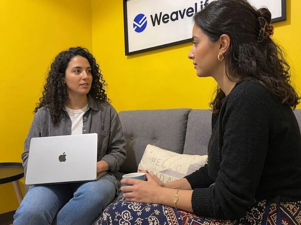 Two people talking on the office sofa under the WeaveLines sign, a laptop open on one of their laps.