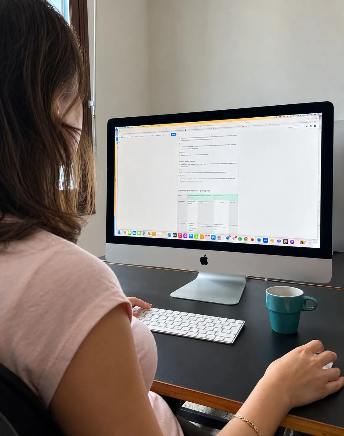 Someone at a desk reading a long document on a large screen, a table of notes under the text and a mug beside the keyboard.
