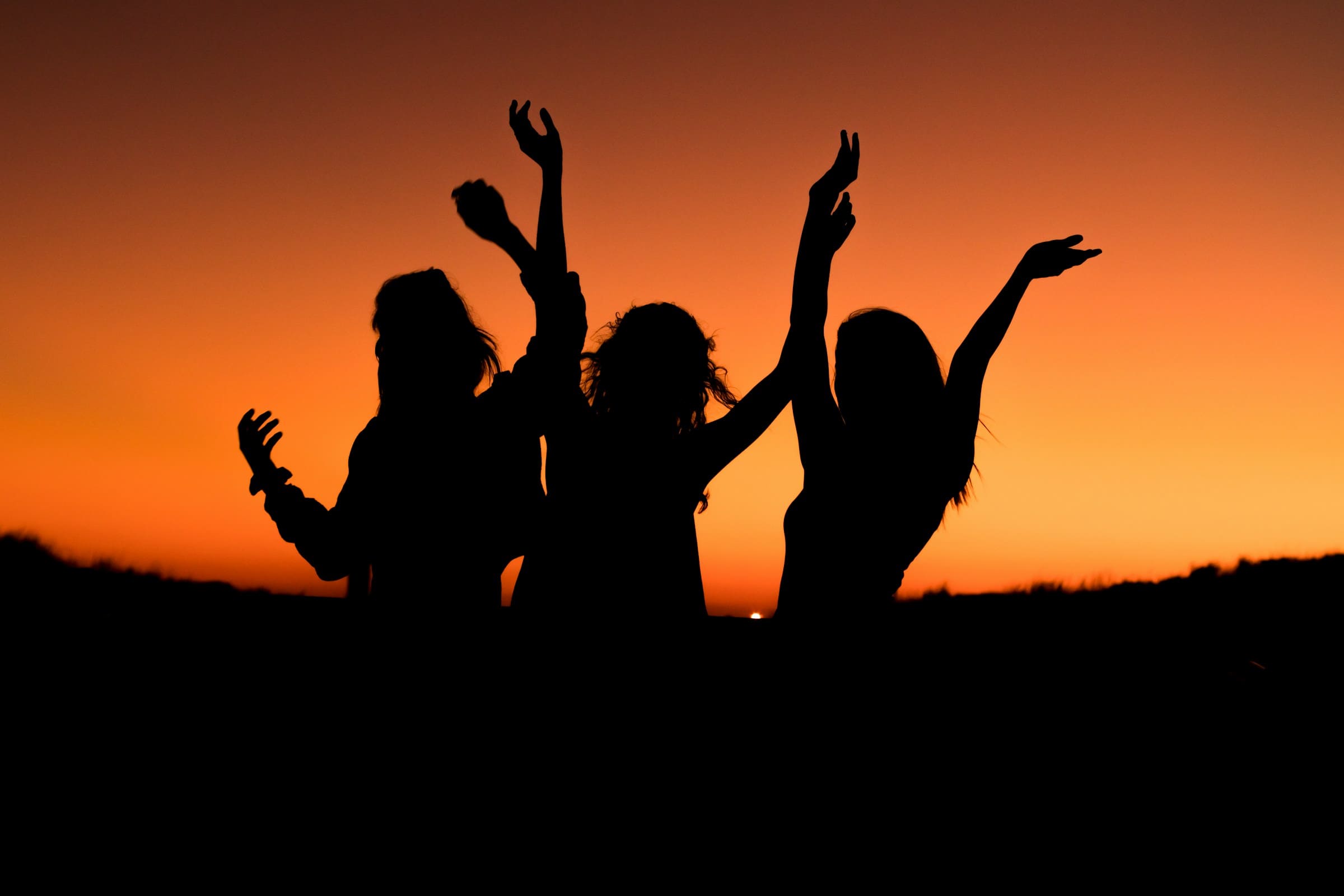 Three friends dancing with raised arms, silhouetted against an orange sunset sky.